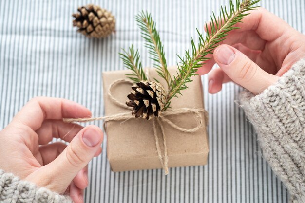 Ein Mädchen in einem Pullover hält ein Geschenk, das mit ihren eigenen Händen verziert ist. Schöne Geschenkbox aus Kraftpapier, Weihnachten oder Neujahr. Geschenk in Frauenhänden, Nahaufnahme.