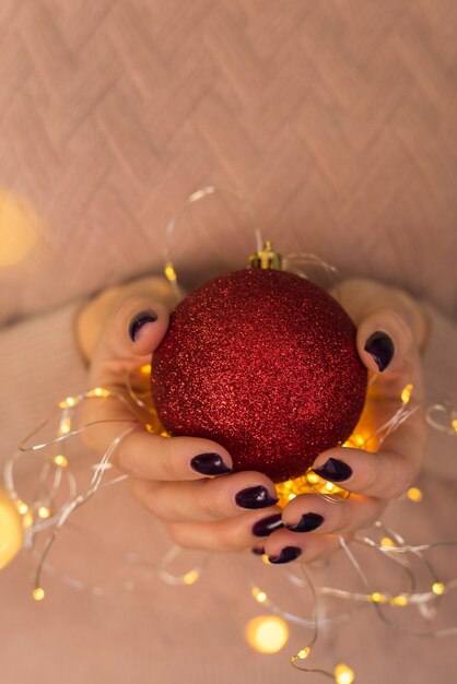 Ein Mädchen in einem rosa Strickjackengriff gibt eine rote glänzende Weihnachtskugel in ihrem hs mit gelben Knoblauchlichtern. Schön getönt.