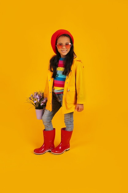 ein Mädchen in einem roten Barett mit rosa Brille in einem gelben Regenmantel mit einem Eimer mit Blumen