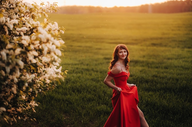 Ein Mädchen in einem roten Kleid mit roten Lippen steht neben einem großen weißen blühenden Baum bei Sonnenuntergang.