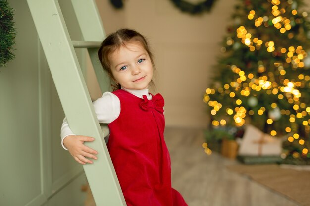 Ein Mädchen in einem roten Kleid steht in einem Raum mit einem Weihnachtsbaum und lächelt süß