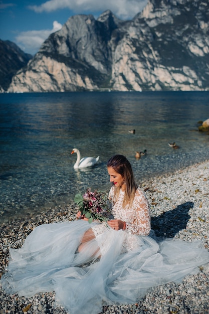 Ein Mädchen in einem schicken weißen Kleid sitzt am Ufer des Gardasees. Eine Frau wird vor dem Hintergrund eines Berges und eines Sees in Italien fotografiert. Torbole.