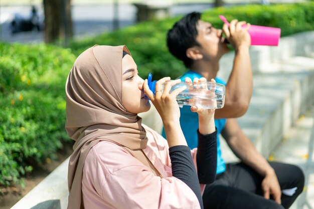 Ein Mädchen in einem Schleier und ein junger Mann sitzen mit einer Flasche trinkend, nachdem sie im Park im Freien Sport treiben