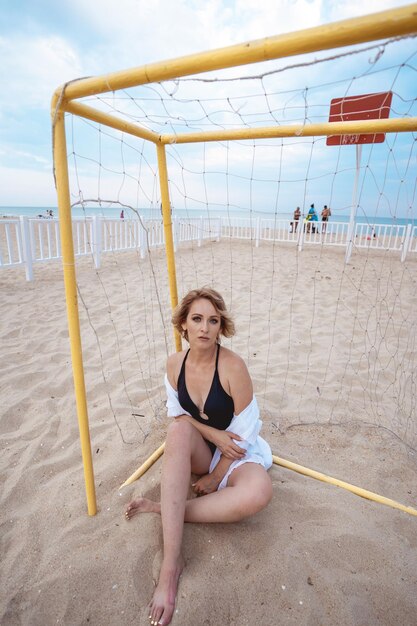 ein mädchen in einem schwarzen badeanzug und einem weißen hemd sitzt am tor zum beachsoccer