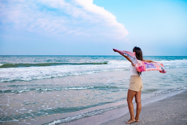 Ein Mädchen in einem sommerlichen T-Shirt und Shorts geht am Strand entlang, blickt auf den Horizont und hält einen Schal in den Händen