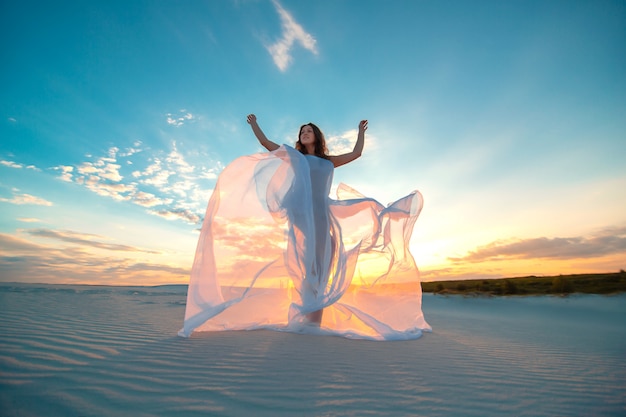 Ein Mädchen in einem weißen Kleid der Fliege tanzt und wirft in der Sandwüste bei Sonnenuntergang auf