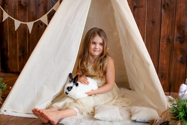 Ein Mädchen in einem weißen Kleid sitzt in einem Zelt Wigwam mit einem Kaninchen, Osterthema