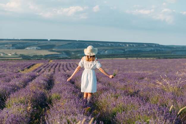 Ein Mädchen in einem weißen Kleid und einem Hut mit Blumensträußen in den Händen steht auf einem Lavendelfeld zurück