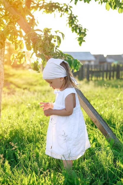 Ein Mädchen in einem weißen Kleid und einem Kopftuch steht unter einem Apfelbaum im Sonnenlicht