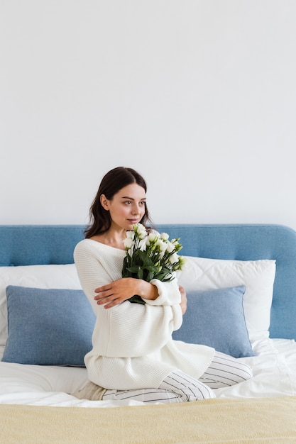 Ein Mädchen in einem weißen Pullover sitzt auf einem Bett und hält einen Strauß weißer Rosen in der Hand