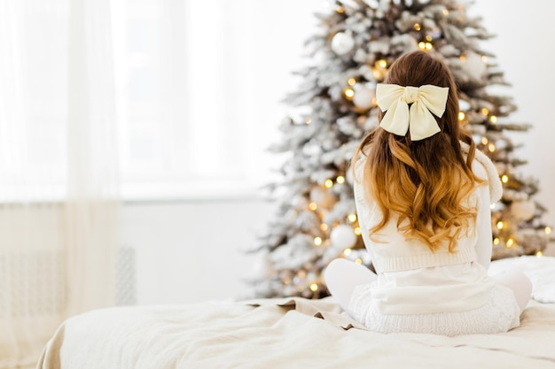 Ein Mädchen in einem weißen Strickkleid mit Haarnadeln in Form einer Schleife sitzt mit dem Rücken zur Kamera auf dem Bett und blickt auf den Weihnachtsbaum