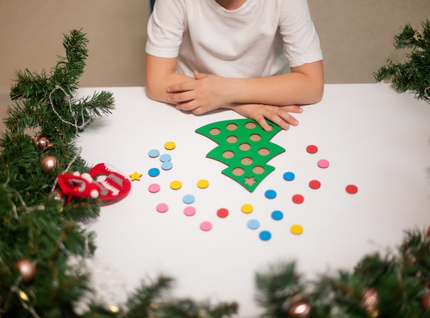 Ein Mädchen in einem weißen T-Shirt mit einer Neujahrsmütze auf dem Kopf schneidet einen Spielzeug-Original-Weihnachtsbaumsorter. Konzept der Weihnachtstraditionen. Entwicklung der Feinmotorik Montessori zu Weihnachten.