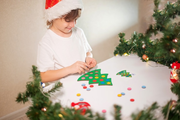 Foto ein mädchen in einem weißen t-shirt mit einer neujahrsmütze auf dem kopf schneidet einen spielzeug-original-weihnachtsbaumsorter. konzept der weihnachtstraditionen. entwicklung der feinmotorik montessori zu weihnachten.