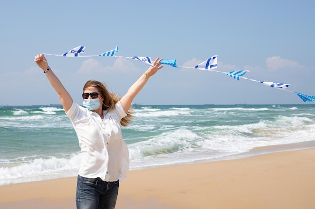 Ein Mädchen in einer medizinischen Maske hält eine winkende Girlande mit israelischen Flaggen an der Mittelmeerküste Unabhängigkeitstag Israel Yom Ha'atzmaut Konzept
