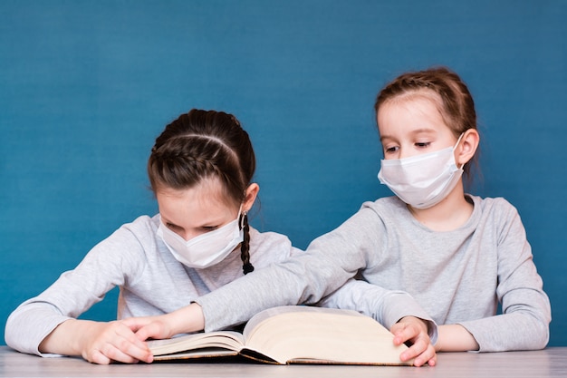 Ein Mädchen in einer medizinischen Maske in Quarantäne liest ein Buch, und ein anderes Mädchen in einer Maske nimmt es ihr ab. Bildung für Kinder isoliert in einer Epidemie