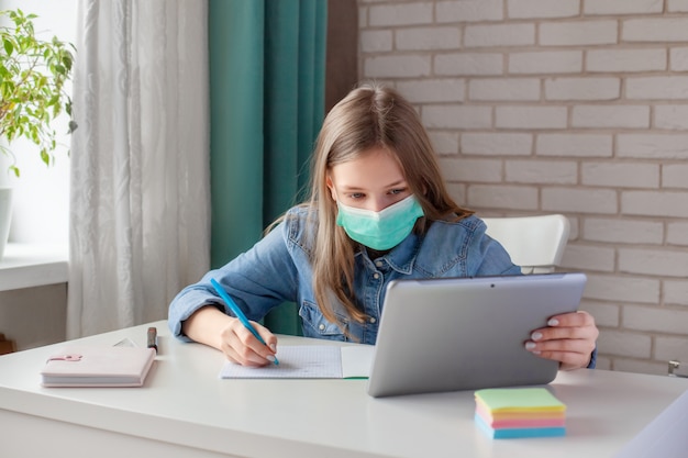 Ein Mädchen in einer medizinischen Maske lernt zu Hause mit einem digitalen Tablet-Laptop und macht Hausaufgaben. Online-Fernunterricht