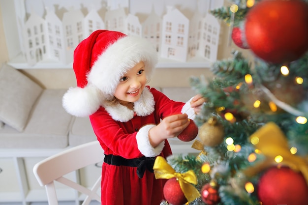 Ein Mädchen in einer Weihnachtsmütze schmückt den Weihnachtsbaum mit Spielzeug und Kugeln und wartet auf ein Geschenk von Santa