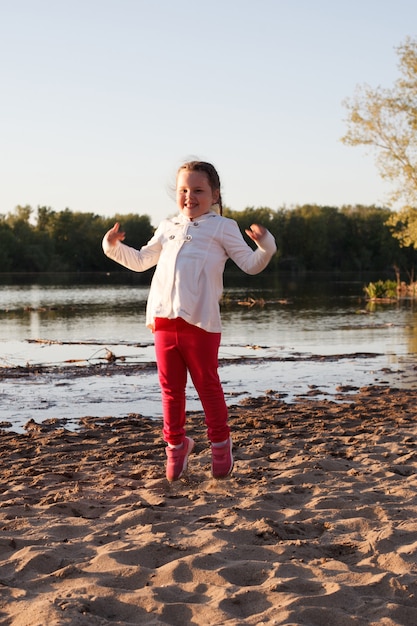 Ein Mädchen in einer weißen Jacke und einer rosa Hose springt auf den Sand am Strand am Flussufer während des Sonnenuntergangs.