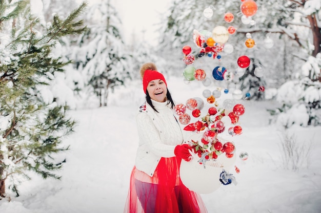 Ein Mädchen in einer weißen Jacke wirft Weihnachtskugeln, um den Weihnachtsbaum zu schmücken. Ein Mädchen wirft Weihnachtsschmuck aus einem Korb in den Winterwald