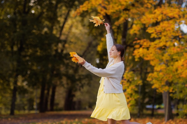 Ein Mädchen in gelber Kleidung in einem Herbstpark freut sich im Herbst und hält gelbe Blätter in ihren Händen