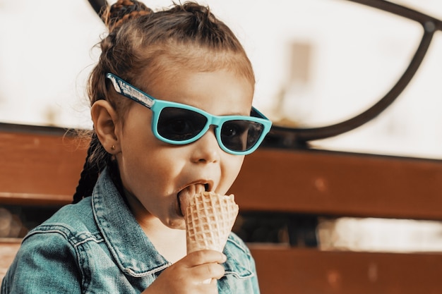 Ein Mädchen in Jeansanzug und Sonnenbrille isst Eis auf einer Parkbank. Foto in hoher Qualität