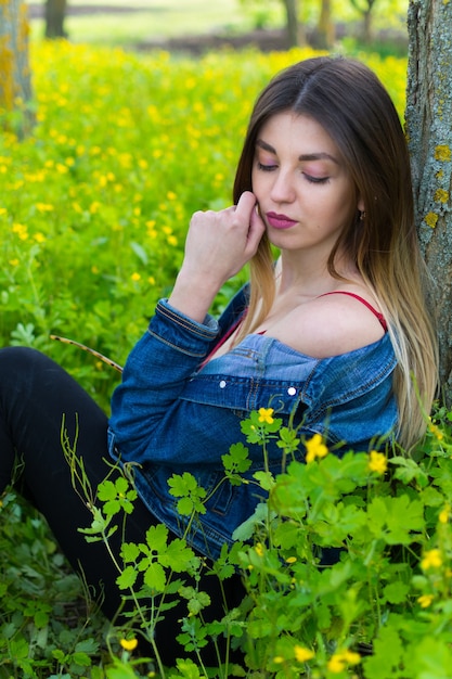 Ein Mädchen in Jeanskleidung mit schönen Haaren sitzt mit einem verträumten Gesichtsausdruck an einem Baum gelehnt.