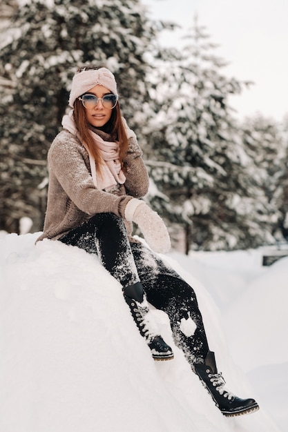 Ein Mädchen in Pullover und Brille sitzt im Winter auf einem schneebedeckten Hintergrund im Wald.