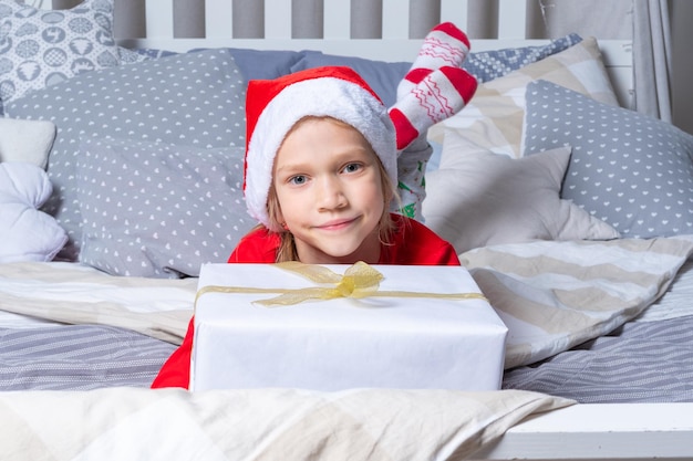 Ein Mädchen in rotem Schlafanzug und Weihnachtsmütze hält eine große Geschenkbox, die auf dem Bett im Kinderzimmer liegt Weihnachts- und Neujahrsgeschenke