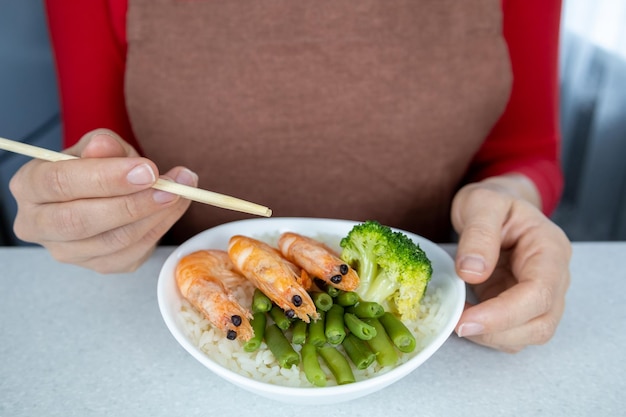 Ein Mädchen in roter Bluse und brauner Schürze hält einen Teller mit goldenem Reis und drei große Riesengarnelen in ihren Händen Das Gericht ist mit grünen Bohnen und Brokkoli dekoriert Asiatisches Lebensmittelkonzept