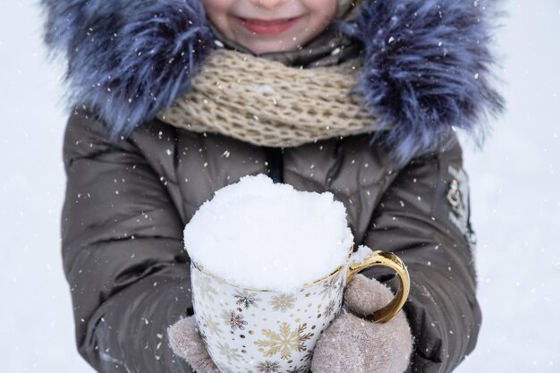 Ein Mädchen in warmer Winter-Outdoor-Kleidung hält einen Becher mit Schnee in den Händen bei kaltem Wetter Saisonalität Weihnachten und Neujahr
