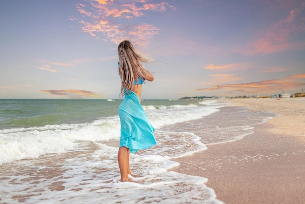 Ein Mädchen mit blonden Haaren in einem bläulichen Badeanzug und einem hellen Schal geht am Strand entlang