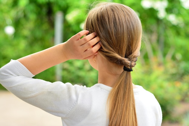 Ein Mädchen mit blonden Haaren und einem weißen Pullover steht rückwärts. Das Mädchen repariert ihr Haar.