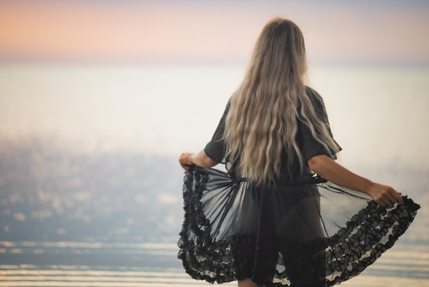 Ein Mädchen mit blonden schicken Haaren in einem schwarzen Bikini und einem Spitzenschal geht die Mündung entlang