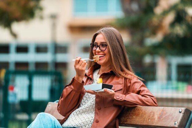 Ein Mädchen mit Brille und modischer Kleidung sitzt auf einer Parkbank und isst köstliche Süßigkeiten. Selektiver Fokus