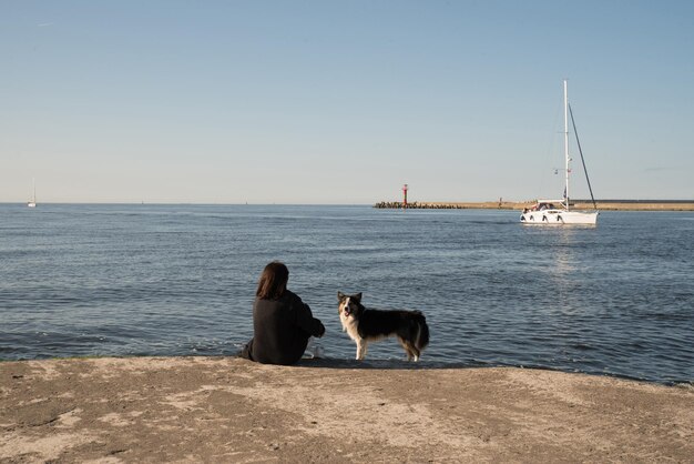 Ein Mädchen mit einem Hund am Ufer, das ein Segelboot betrachtet