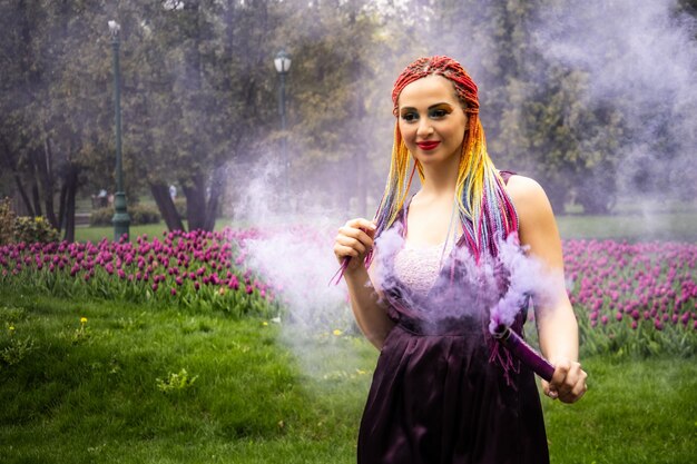 Ein Mädchen mit einem Lächeln mit hellem Make-up und farbigen Zöpfen in einem lila Kleid bläst künstlichen lila Rauch in einem frühlingsblühenden Park