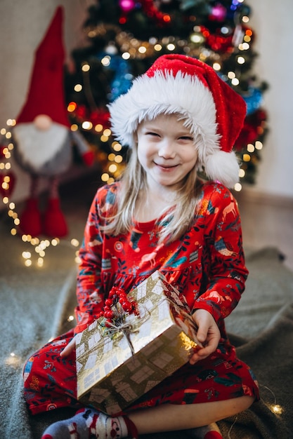 Ein Mädchen mit einem roten Weihnachtsmannhut sortiert Geschenke vor dem Hintergrund eines Weihnachtsbaumes aus