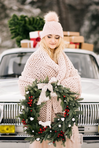 Ein Mädchen mit einem Weihnachtskranz in den Händen bedeckt mit einer Decke und steht auf dem Hintergrund eines Retro-Autos, dessen Dach ein Weihnachtsbaum und Geschenke im schneebedeckten Winterwald ist.