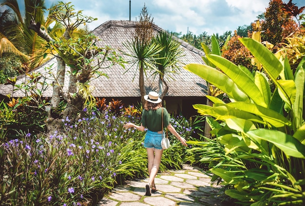 Ein Mädchen mit Hut geht an einem sonnigen Tag durch das Gebiet eines Luxushotels in Ubud. Eine junge Frau geht auf einem Weg, der von leuchtenden Blumen und tropischen Pflanzen umgeben ist, Blick von hinten, Bali, Ubud.