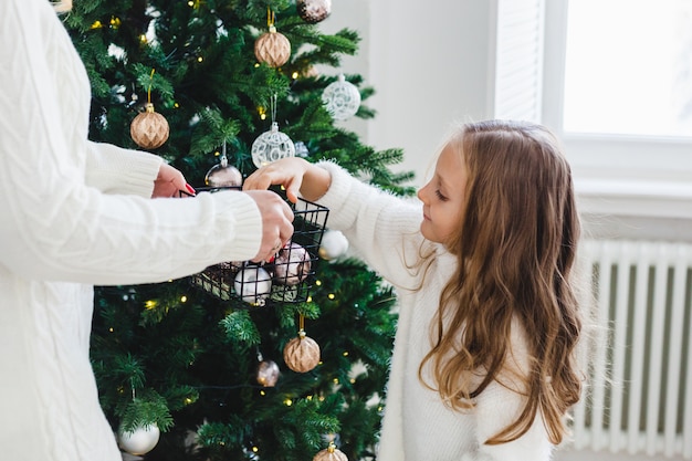 Ein Mädchen mit ihrer Mutter schmücken den Weihnachtsbaum, der Innenraum ist für das neue Jahr und Weihnachten, Familie und Freude, Traditionen geschmückt.