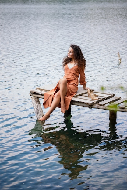 Ein Mädchen mit langen gewellten Locken in einem langen Guipure-Kleid barfuß im Sommer in einem Wald an einem See bei Sonnenuntergang, das auf einem Pantone auf einer hölzernen Pierbrücke steht. Sommer sonniger Tag