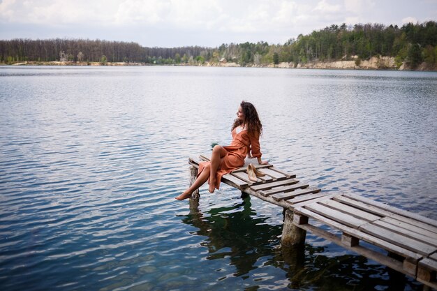 Ein Mädchen mit langen gewellten Locken in einem langen Guipure-Kleid barfuß im Sommer in einem Wald an einem See bei Sonnenuntergang, das auf einem Pantone auf einer hölzernen Pierbrücke steht. Sommer sonniger Tag