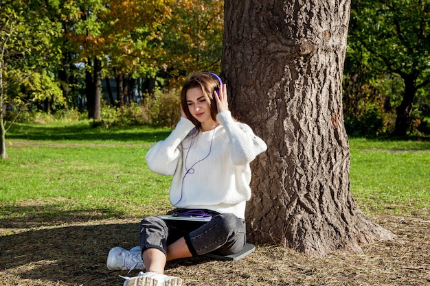 Ein Mädchen mit langen Haaren in einem weißen Pullover und Jeans hört im Park sitzend ihre Lieblingsmusik