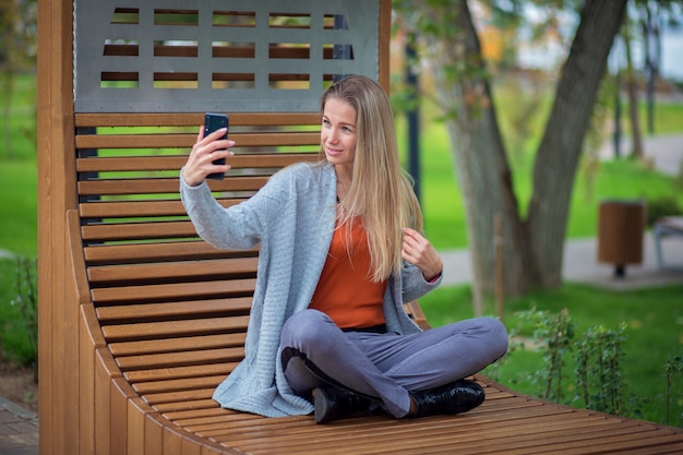 Ein Mädchen mit langen Haaren und einer grauen Jacke sitzt auf einer Bank im Park und macht ein Selfie auf einem Smartphone