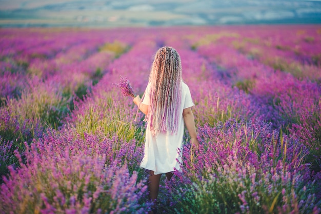Ein Mädchen mit lila Zöpfen auf dem Kopf, das ein weißes Kleid und einen Lavendelstrauß in den Händen trägt