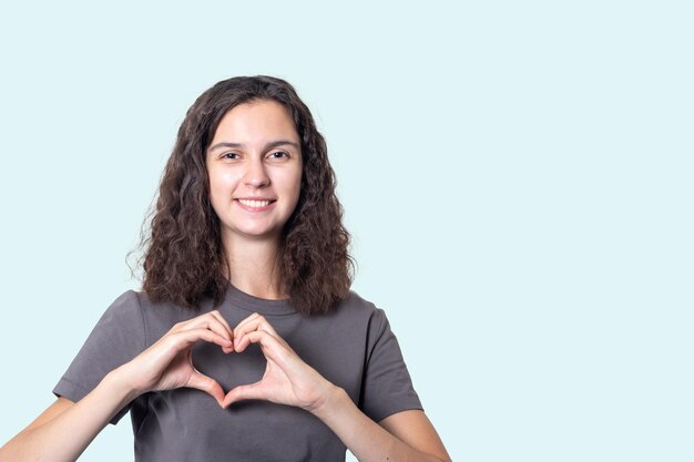 Ein Mädchen mit lockigem Haar macht Hände in Form eines Herzens auf hellblauem Hintergrund. Herzkrankenversicherung, Spende, glücklicher Wohltätigkeitsfreiwilliger, Weltherztag, Welttag der psychischen Gesundheit, Krebstag