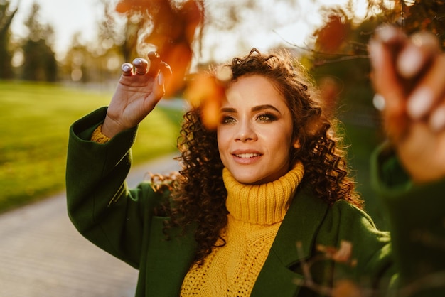 Ein Mädchen mit lockigen langen Haaren in einem leuchtend gelben Pullover berührt mit ihren Händen Herbstblätter auf Branc