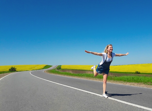 Ein Mädchen mit offenen Armen in Form eines fliegenden Vogels steht auf einer breiten Asphaltstraße, die sich perspektivisch schlängelt, mitten auf einem Feld