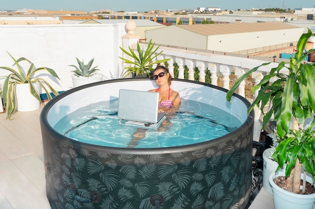 Ein Mädchen mit Sonnenbrille im Wasser eines aufblasbaren Mini-Pools arbeitet auf einem Laptop-Computer das Konzept