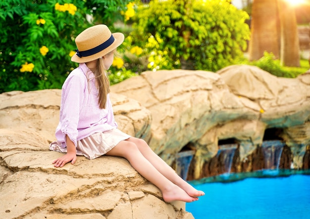 Ein Mädchen mit Strohhut sitzt mit abgewandtem Gesicht am Pool zwischen tropischen Pflanzen in der Sonne...